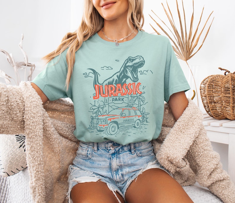 Pu&ograve; includere: Una t-shirt verde chiaro con una grafica verde scuro raffigurante un dinosauro T-Rex, una scena nella giungla e un veicolo. Il testo JURASSIC PARK &egrave; stampato in arancione e rosso. La maglietta ha maniche corte e girocollo, indossata con pantaloncini di jeans.