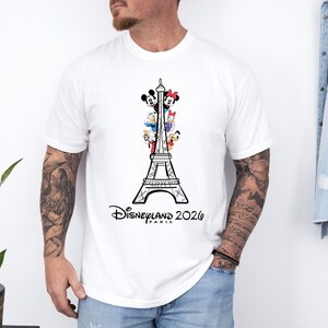 May include: A white short-sleeved t-shirt featuring a black line drawing of the Eiffel Tower. The tower is decorated with colorful cartoon characters including Mickey Mouse, Minnie Mouse, Donald Duck, Daisy Duck, Goofy, and Pluto. Text reads Disneyland Paris 2026.