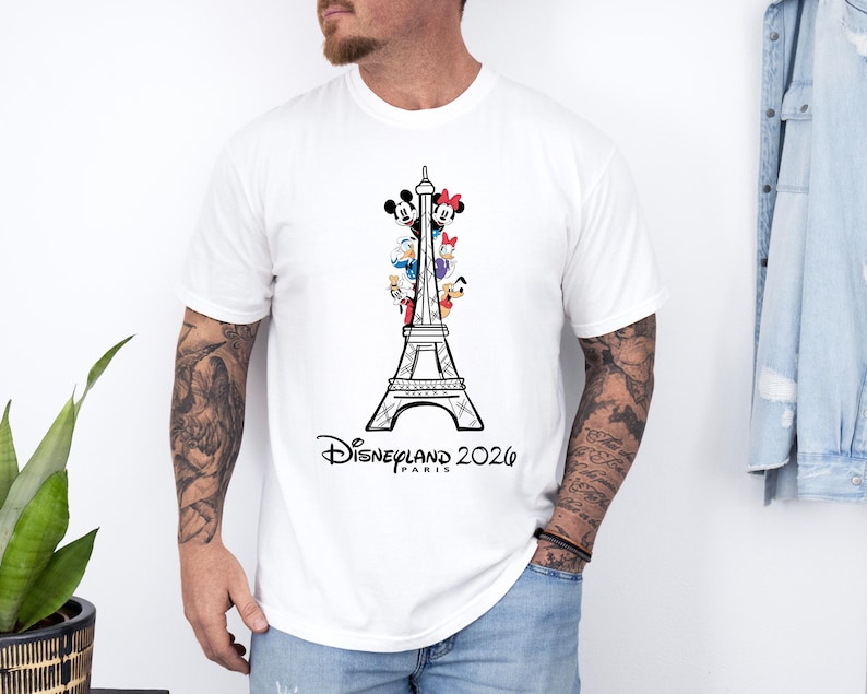 May include: A white short-sleeved t-shirt featuring a black line drawing of the Eiffel Tower. The tower is decorated with colorful cartoon characters including Mickey Mouse, Minnie Mouse, Donald Duck, Daisy Duck, Goofy, and Pluto. Text reads Disneyland Paris 2026.
