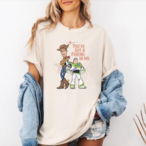 May include: A cream-coloured short-sleeve t-shirt featuring a graphic print of Woody and Buzz Lightyear standing side by side. The text "You've Got A Friend In Me" is printed in a brown font next to the characters, accompanied by a star and atom icon.
