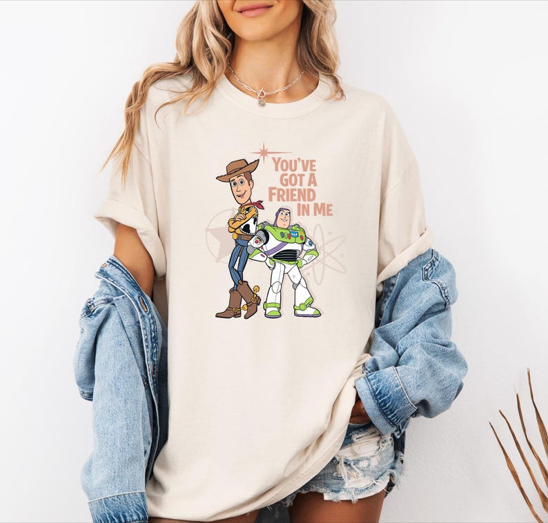 May include: A cream-coloured short-sleeve t-shirt featuring a graphic print of Woody and Buzz Lightyear standing side by side. The text "You've Got A Friend In Me" is printed in a brown font next to the characters, accompanied by a star and atom icon.