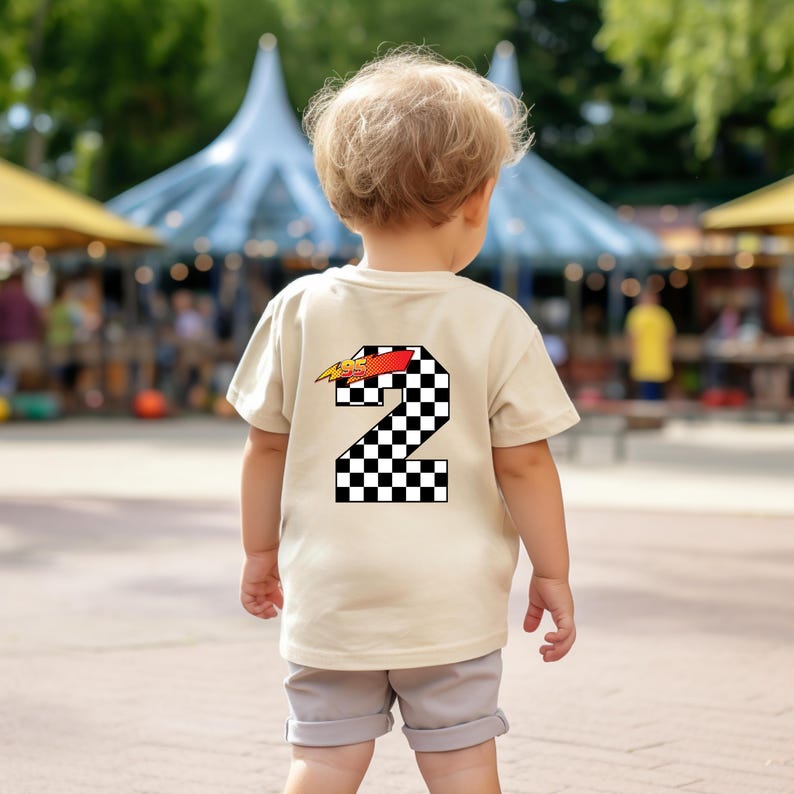 May include: A beige short-sleeve t-shirt featuring a large number two on the back. The number is filled with a black and white checkered pattern and topped with a red and yellow lightning bolt graphic. The shirt is paired with light gray shorts.