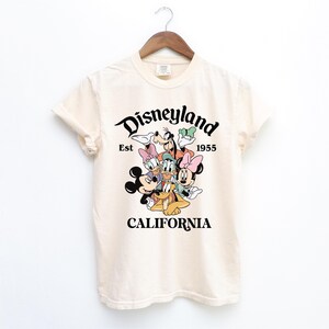May include: A cream-colored t-shirt with a black graphic of Mickey Mouse and friends. The graphic says "Disneyland Est 1955 California".