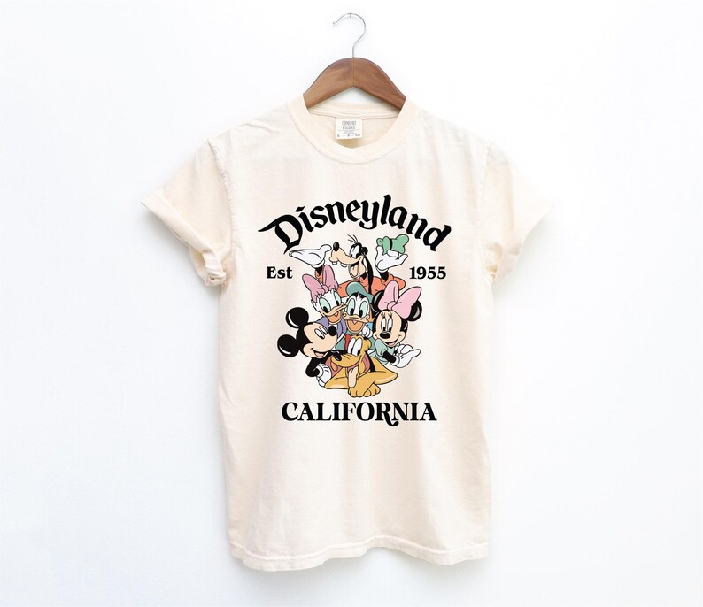 May include: A cream-colored t-shirt with a black graphic of Mickey Mouse and friends. The graphic says "Disneyland Est 1955 California".