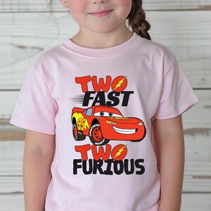 May include: A light pink short-sleeved t-shirt featuring a graphic of the red race car Lightning McQueen. The text reads TWO FAST TWO FURIOUS with yellow lightning bolt accents replacing the letter O in both words, printed in black and red.