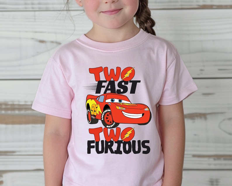 May include: A light pink short-sleeved t-shirt featuring a graphic of the red race car Lightning McQueen. The text reads TWO FAST TWO FURIOUS with yellow lightning bolt accents replacing the letter O in both words, printed in black and red.