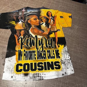 Op de afbeelding: Wit T-shirt met een zwart en geel ontwerp met een groep dansers en de tekst "Kemya My Favorite Dancer Calls Me Cousins".
