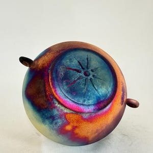 May include: A round ceramic bowl with two small side handles. The exterior features a vibrant, iridescent glaze with blended hues of orange, magenta, deep blue, and yellow. The center of the bowl displays a raised, star-shaped sunburst design.
