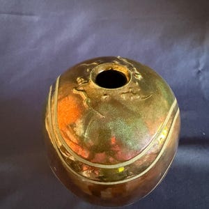 May include: A spherical ceramic vase with a metallic, iridescent glaze featuring shifting tones of bronze, copper, green, and gold. The surface is accented with thin, horizontal, light-colored lines and a small, circular opening at the top.