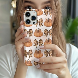 May include: A smartphone case featuring a repeating pattern of orange pumpkins and leopard print bows on a light background. The design includes multiple pumpkins with stems and bows arranged in a grid-like layout across the entire surface.