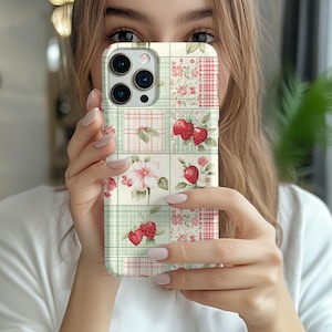 May include: A phone case with a patchwork design featuring strawberries, flowers, and plaid patterns in red, pink, and green. The case has a square grid pattern and a camera cutout.