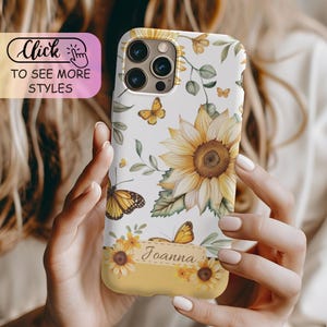 May include: A white smartphone case featuring a pattern of yellow sunflowers, green leaves, and orange butterflies. A rectangular label at the bottom contains the name Joanna in cursive script. The case has a cutout for a triple camera system.