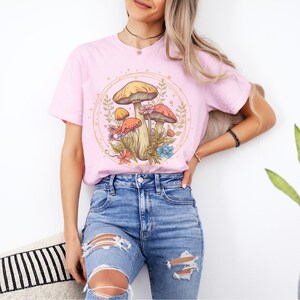 以下が含まれることがあります： 金色の円の中に、カラフルなキノコと花が描かれたピンクのTシャツ。キノコは茶色の茎と赤と黄色の帽子をしています。