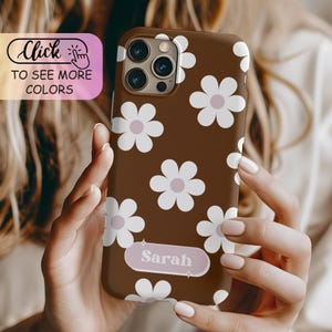 May include: A brown smartphone case featuring a repeating pattern of white flowers with light pink centers. A horizontal rectangular label with rounded corners is centered near the bottom, displaying the name Sarah in white serif font.