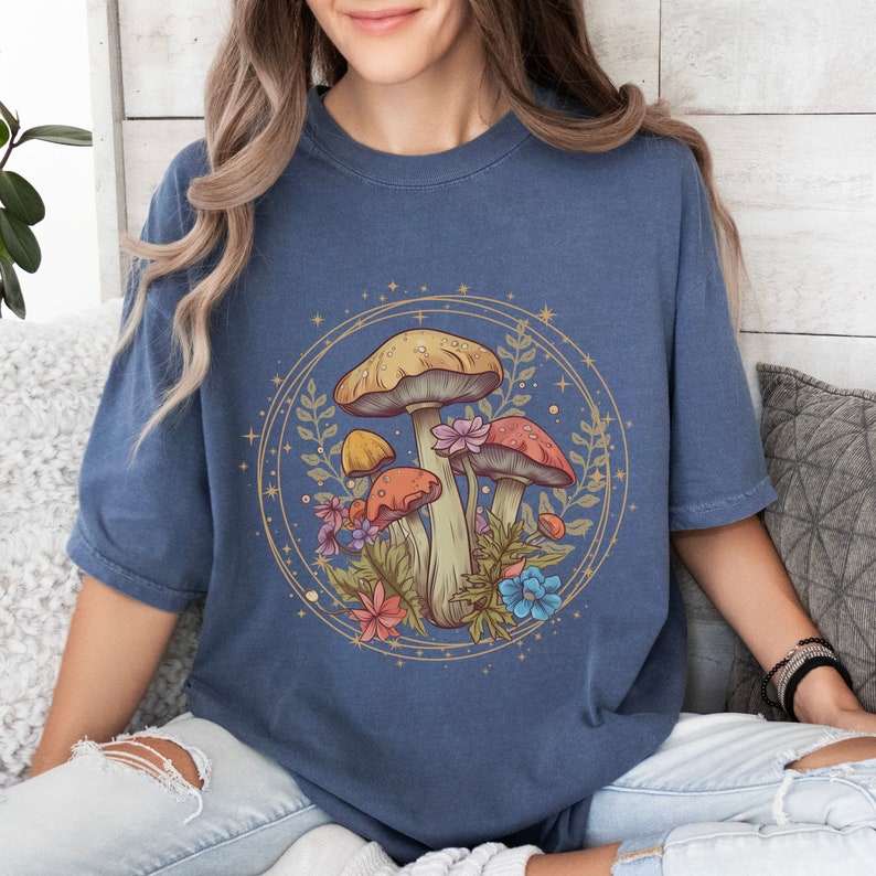 以下が含まれることがあります： 円の中にキノコと花が描かれた青いTシャツ。キノコは赤、オレンジ、黄色。花はピンク、青、黄色です。