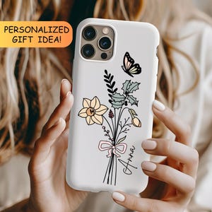 May include: A white smartphone case featuring a line art illustration of a bouquet with a yellow daffodil, green holly leaves, and a small butterfly. A pink ribbon is tied around the stems, and the name Anna is written in black script.