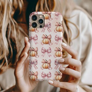May include: A smartphone case featuring a repeating pattern of orange pumpkins and light pink bows. The bows display various textures including gingham, polka dots, and solid patterns, set against a soft, light-colored background.
