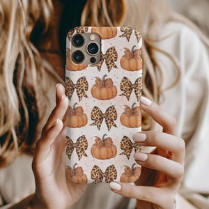 May include: A smartphone case featuring a repeating pattern of orange pumpkins and leopard print bows on a light cream background. The design includes multiple pumpkins with stems and bows arranged in a grid, with a camera cutout at the top left.