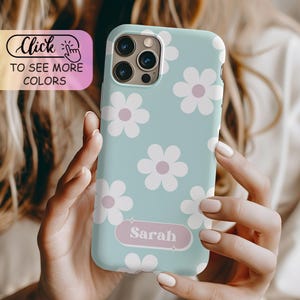 May include: A light blue phone case featuring a repeating pattern of white flowers with light purple centers. A horizontal light purple rectangular label with rounded corners is centered near the bottom, displaying the name Sarah in white text.