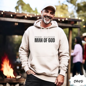 May include: A sand-colored hoodie with the text "PRIMARY OCCUPATION MAN OF GOD" in black block letters. The hoodie has a front pocket and a drawstring hood. The image shows a person wearing the hoodie.