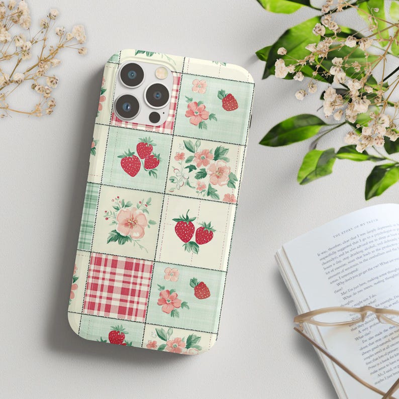 May include: A white phone case with a patchwork design featuring red strawberries, pink flowers, and green leaves on a light green background.