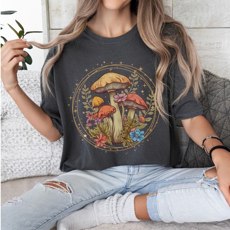 以下が含まれることがあります： 円の中にキノコと花が描かれたダークグレーのTシャツ。キノコは黄色、オレンジ、赤。花はピンク、青、黄色です。