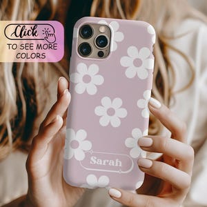 May include: A mauve phone case featuring a repeating pattern of white six-petaled flowers. The case includes a rectangular label at the bottom with the name Sarah printed in white text. The camera cutout is visible at the top left corner.