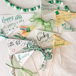 Pode incluir: Uma coleção de bandeiras de bandeirolas do Dia de São Patrício com frases como "Happy st. patty's day" e "my lucky charm". As bandeiras são brancas e amarelas com detalhes verdes, trevos e laços. Uma grinalda de contas verdes está no fundo.