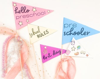 Op de afbeelding: Vier kleurrijke driehoekige vlaggen met verschillende berichten voor een kleuterschool of eerste schooldag viering. De vlaggen zeggen "hallo kleuterschool", "schoolregels", "eerste dag" en "kleuter".