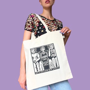 Könnte beinhalten: Eine weiße Einkaufstasche mit einer Schwarzweißillustration von drei Personen, die in einer Reihe sitzen. Der Text "LET'S RECLAIM OUR SPACE!" ist unter der Illustration gedruckt.