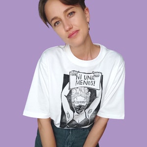May include: White t-shirt with a black and white illustration of a woman wearing a face mask and holding a sign that says "NI UNA MENOS!"