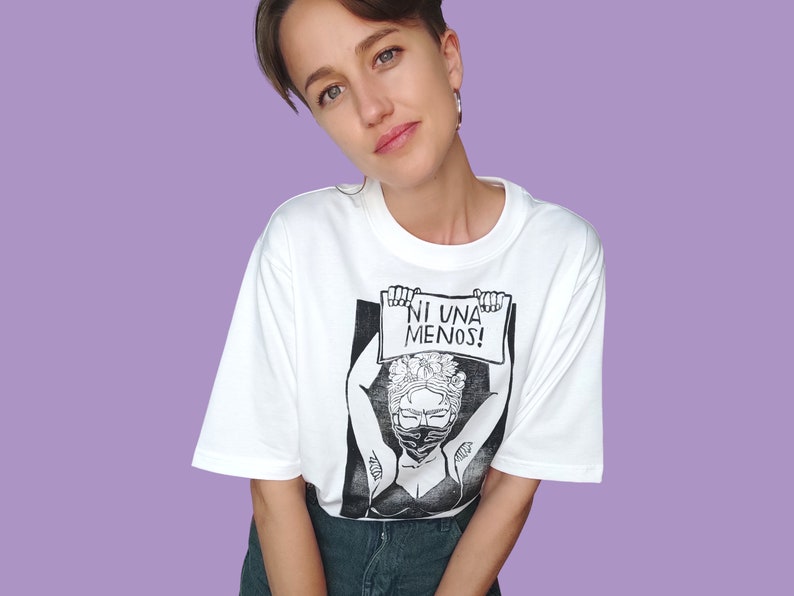 May include: White t-shirt with a black and white illustration of a woman wearing a face mask and holding a sign that says "NI UNA MENOS!"
