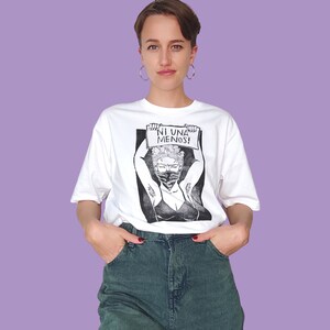 May include: White t-shirt with a black and white illustration of a woman wearing a face mask and holding a sign that says "NI UNA MENOS!"