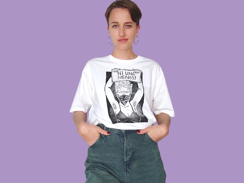 May include: White t-shirt with a black and white illustration of a woman wearing a face mask and holding a sign that says "NI UNA MENOS!"