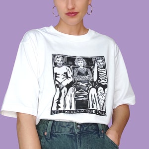 Könnte beinhalten: Weißes T-Shirt mit einem schwarz-weißen Grafikdruck von drei Personen, die in einem Raum sitzen. Der Text "Let's Reclaim Our Space" ist unter dem Grafikdruck gedruckt.