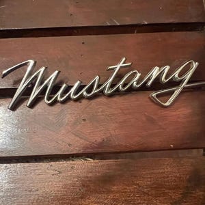 May include: A chrome-colored "Mustang" emblem, in a cursive script, is displayed on a wooden surface. The emblem is a classic car part, likely for a Ford Mustang. The wooden surface has a dark brown color and a wood grain texture.