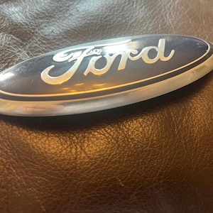 May include: Oval-shaped Ford emblem with a dark blue background and silver lettering. The word "Ford" is written in a stylized script. The emblem has a silver border and is set against a brown leather surface.