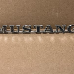 May include: A silver "MUSTANG" emblem, likely for a car, is displayed against a neutral brown background. The letters are in a bold, capitalized font, and the emblem appears to be made of metal.