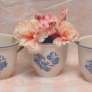May include: Three off-white ceramic flower pots with blue floral designs and blue rims. The center pot holds pink and peach flowers. The pots are arranged on a light pink surface.