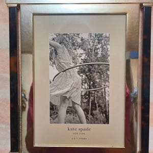 May include: A rectangular Kate Spade picture frame with a gold-colored metal border and black and brown accents. The frame holds a black and white photo of a person with a hula hoop. The text "KATE SPADE NEW YORK" and "2 X 7 FRAME" are visible.