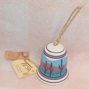 May include: A hand-painted ceramic bell featuring a desert landscape with cacti in shades of blue, pink, and purple. The bell has a braided tan cord for hanging and a beige paper tag attached that reads TESA Hand Painted by Maack Co.