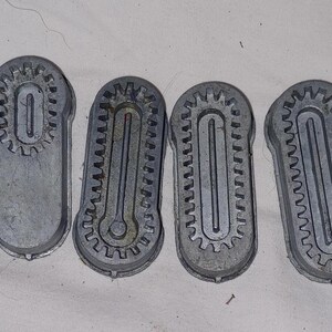 May include: Four vintage, cast-aluminum molds with a gear-like design around the edges. Each mold has an oval shape with a central, elongated depression. The molds are a dull silver color and appear to be well-used.