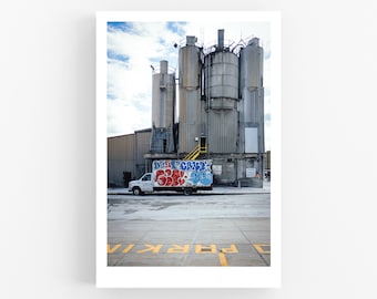 Può includere: Un camioncino bianco parcheggiato davanti a un grande edificio industriale con diversi silos alti. I silos sono dipinti di grigio e hanno un design di graffiti blu e rosso su un lato. Il camioncino ha un logo aziendale sul lato e le parole "Park" sono dipinte a terra davanti al camioncino.