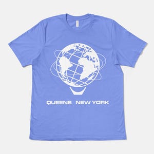 以下が含まれることがあります： ペリウィンクルブルーの半袖Tシャツ。軌道線に囲まれた地球儀の白いグラフィックが中央に配置されています。地球儀の下には、太字の白いサンセリフ体で「QUEENS NEW YORK」という文字がプリントされています。Tシャツはクルーネック仕様です。