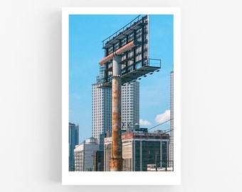Può includere: Un grande cartellone pubblicitario arrugginito si erge alto sullo sfondo di un cielo blu e di edifici cittadini. Il cartellone è vuoto e sembra in stato di abbandono.
