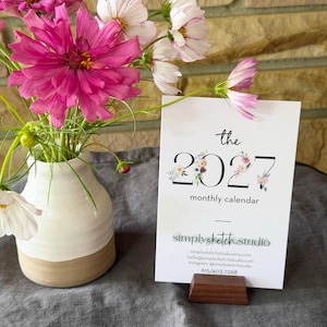 May include: A white ceramic vase with a tan base holds pink and white cosmos flowers next to a white card on a dark wood stand. The card reads: the 2027 monthly calendar, simplysketchstudio, simplysketchstudio.etsy.com, hello@simplysketchstudio.com, instagram @simplysketchstudio, 615/403.7268.