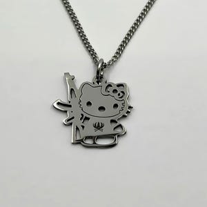 Puede incluir: Collar plateado con un colgante con un diseño de gato de dibujos animados estilizado. El gato sostiene un arma. El collar tiene una cadena de eslabones y un cierre de mosquetón. El colgante mide aproximadamente 2,5 cm de altura.