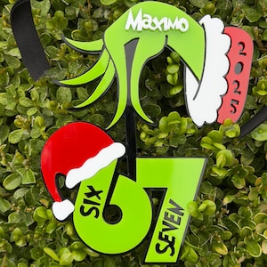 May include: A Christmas ornament with a green hand design, the name "Maximo", and the year "2025". It includes a red and white Santa hat and the numbers "67" with the words "SIX" and "SEVEN". A black ribbon is attached for hanging.