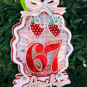 Op de afbeelding: Een gelaagde taarttopper met een roze strik, twee rode aardbeien en het nummer 67 in rood. De naam "Amelia" is in gouden letters geschreven. De topper is versierd met witte en gouden accenten.