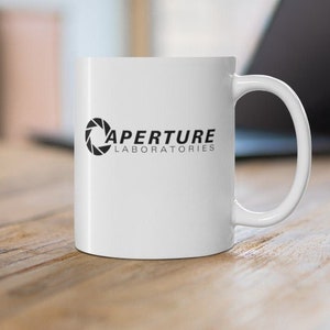 Puede incluir: Taza de café de cerámica blanca con un logotipo negro de Aperture Laboratories que presenta una lente de cámara estilizada. El texto "Aperture Laboratories" está impreso debajo del logotipo.